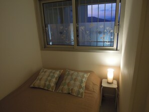 Un dormitori amb un llit, dos coixins, una tauleta de nit amb una làmpada, i una finestra que mostra vistes a les muntanyes.