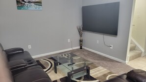 Una sala de estar con un televisor de pantalla plana grande montado en la pared, un sofá de cuero oscuro, una mesa de centro de vidrio y un jarrón decorativo con ramas.