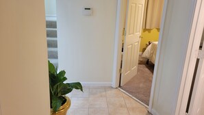 Un pasillo con una planta en maceta, un termostato y una puerta que da a una habitación con una cama y pared amarilla.