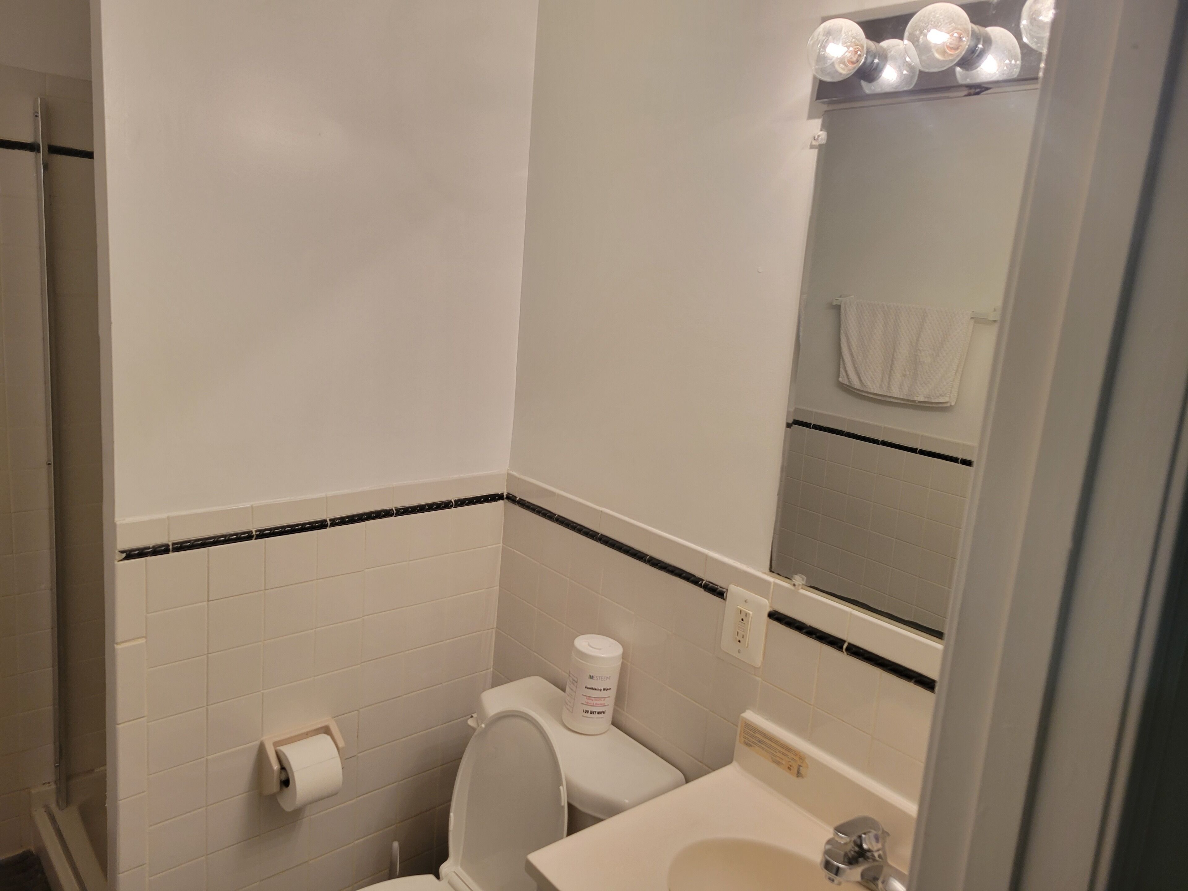 A bathroom with white tiles, a toilet, a sink, and a mirror with lights.