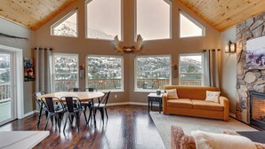 A spacious living room with a wooden ceiling, large windows offering mountain views, a fireplace, and a cozy seating area. - Stunning Alaskan Getaway w/ River & Mtn Views (Eagle River)