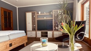 A modern hotel room with a bed, a TV, a desk with a chair, a vase with flowers, and a large window. - mein.guesthouse - apartment 1 - approx. 45sqm with large sun terrace  (Haundorf)