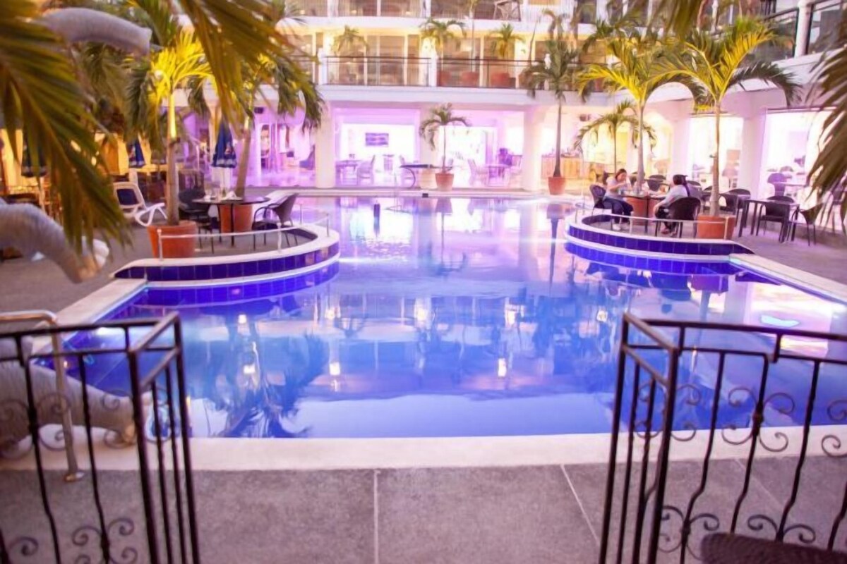 An indoor swimming pool with a circular design, surrounded by lounge areas and palm trees.