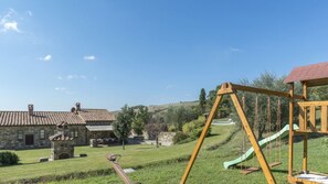 A stone building with a tiled roof, a swing set, and a green slide in a grassy area.