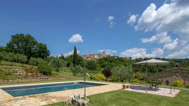 Una piscina con bordo in pietra, circondata da prato e alberi. Sullo sfondo, si vede una collina con un edificio in cima.