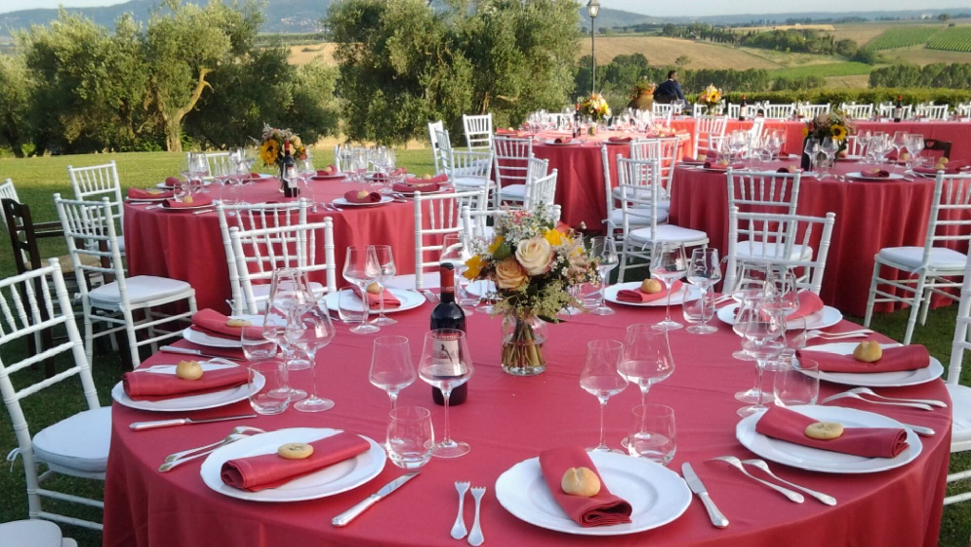 Un espace de restauration en plein air avec des tables rondes recouvertes de nappes rouges, des chaises blanches et des tables soigneusement dressées avec des verres à vin, des assiettes et des couverts.
