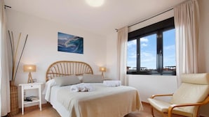Quarto com cama, luminárias de cabeceira, uma cadeira, janela com vista e um quadro na parede.