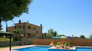 A swimming pool with lounge chairs and umbrellas, surrounded by a stone building with a tiled roof.