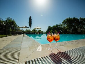 Blick über den Pool mit zwei Gläsern eines Getränks, blauem Himmel und einem Baum in der Ferne.