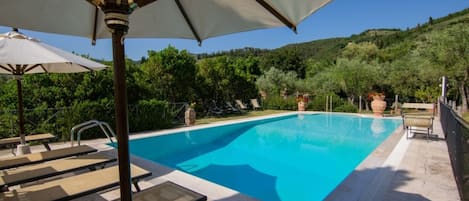 Una piscina con lettini e ombrelloni, immersa nel verde e circondata da colline.