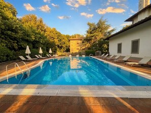 A swimming pool with lounge chairs and umbrellas, surrounded by trees and a building.