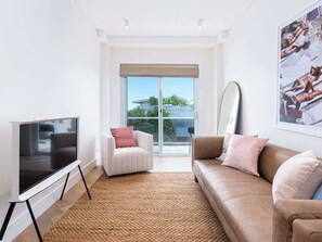 A modern living room with a flat-screen TV, a sofa, a chair, and a large window with a view of greenery.