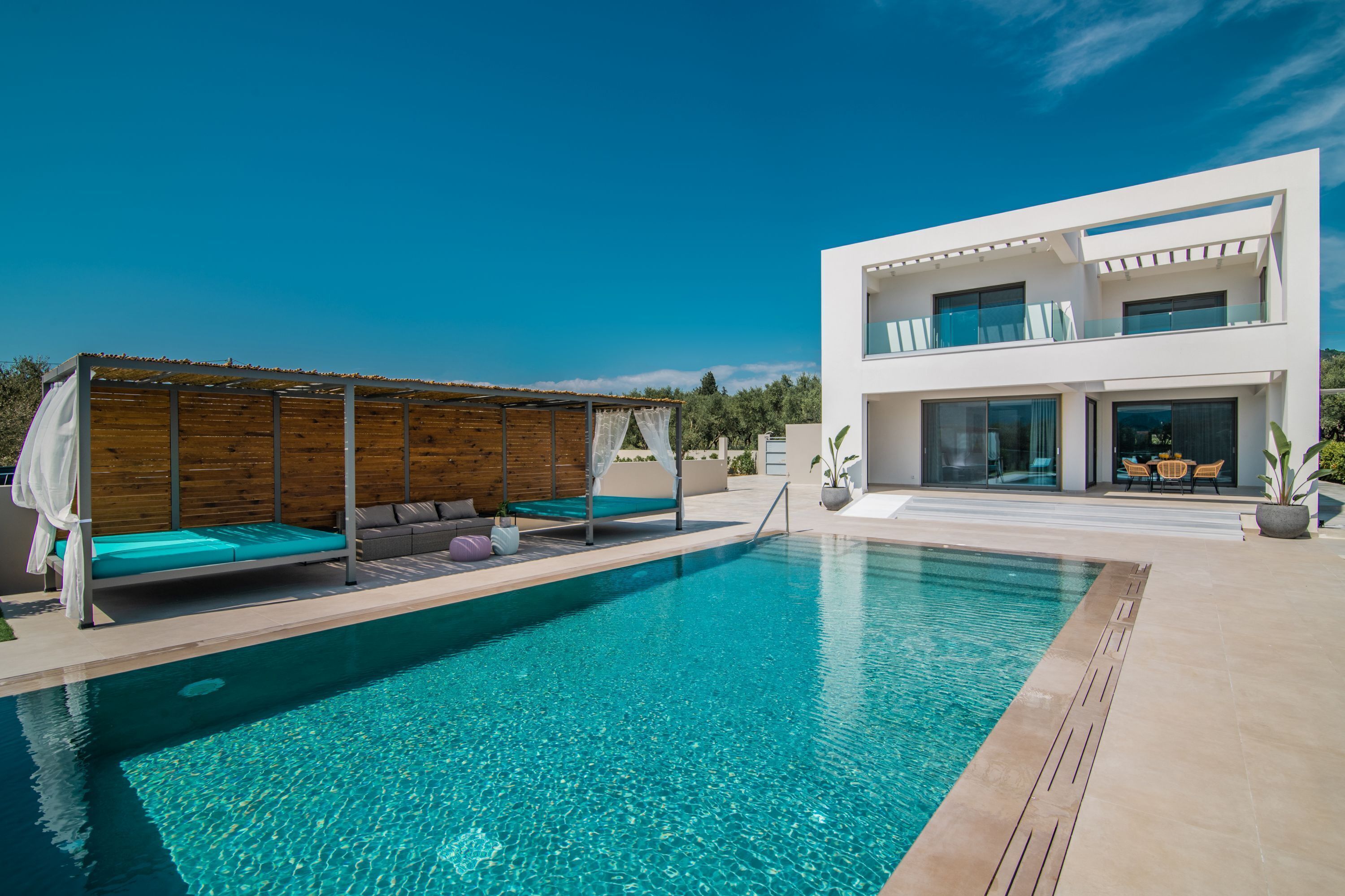 Un espace piscine moderne avec un pavillon en bois, un bâtiment blanc doté de balcons et un ciel d’un bleu limpide.