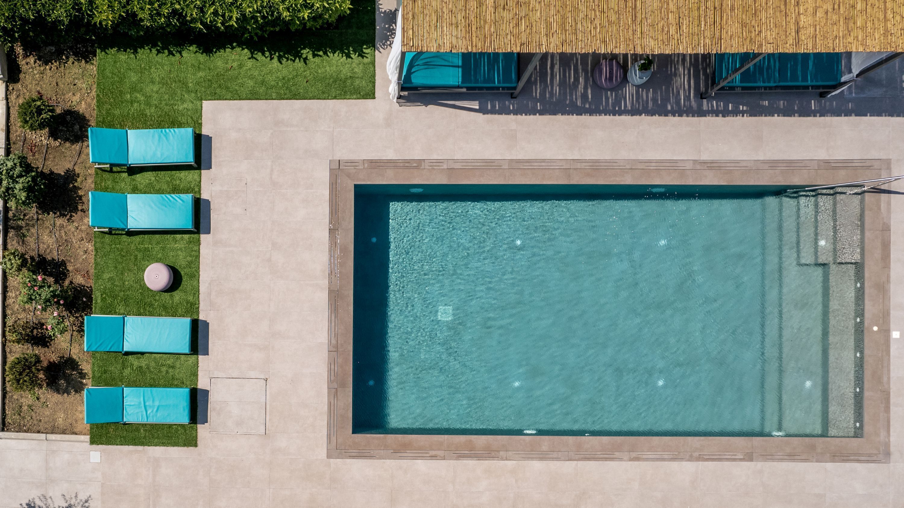 Eine Vogelperspektive zeigt einen rechteckigen Swimmingpool mit einer gefliesten Liegefläche, umgeben von vier blauen Liegestühlen und einem kleinen runden Tisch.