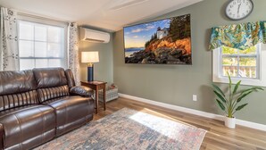 A living room with a brown leather sofa, a patterned rug, a wall-mounted clock, and a large scenic painting. - DT Bar Harbor,2 King beds & Decks, Parks 3 small cars, Town Pickleball Ct @ Back (Bar Harbor)