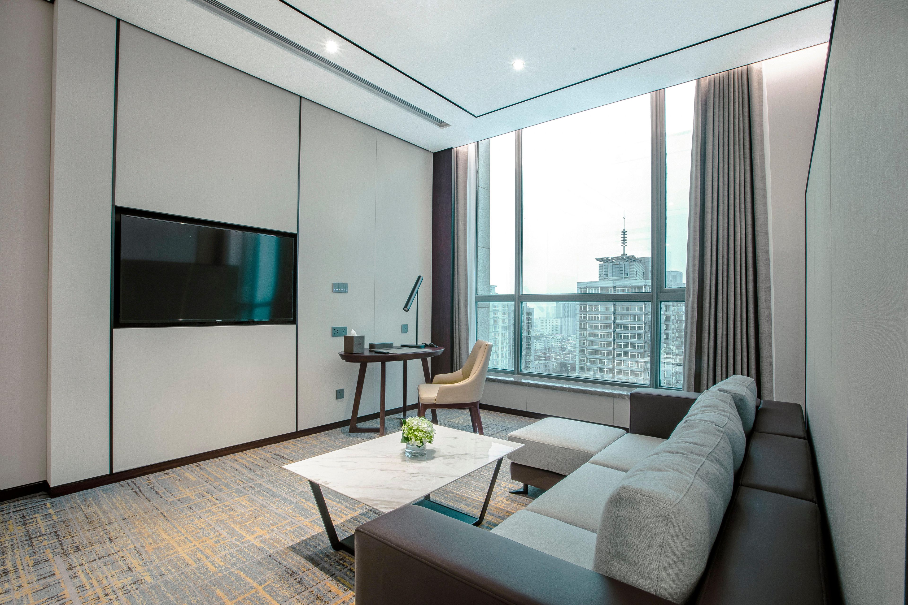 A modern hotel room with a large window, a flat-screen TV, a sofa, a coffee table, and a small desk with a chair.