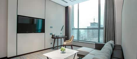 A modern hotel room with a large window, a flat-screen TV, a sofa, a coffee table, and a small desk with a chair.