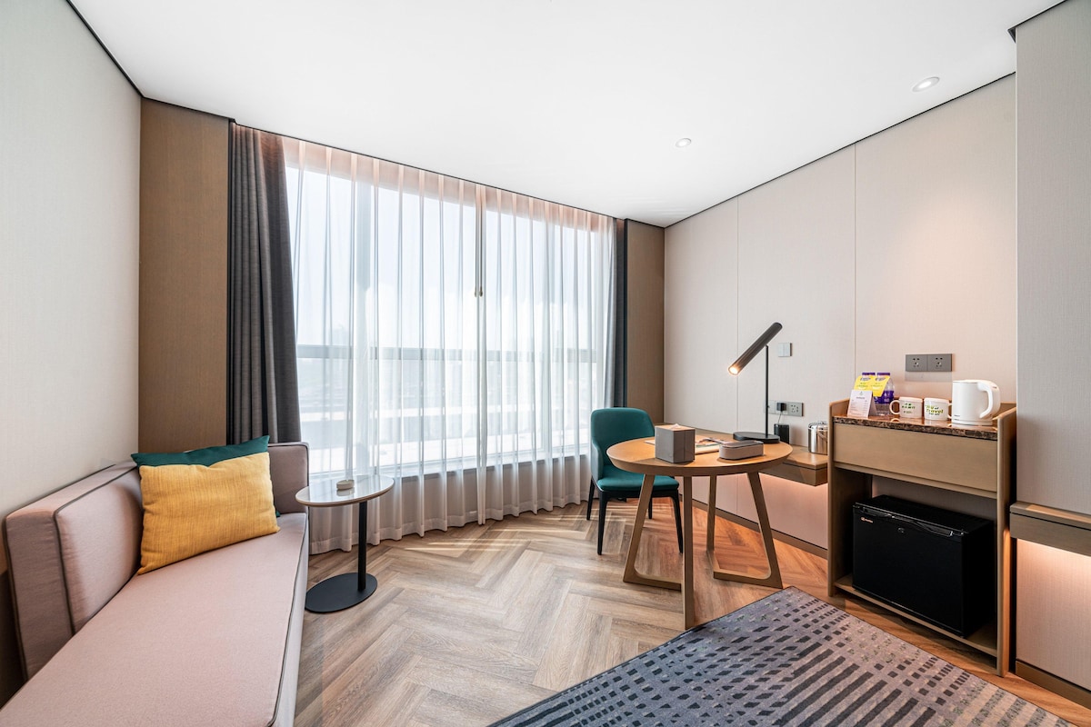A modern hotel room with a sofa, a small table, a chair, a desk, a lamp, a coffee machine, and a mini fridge.