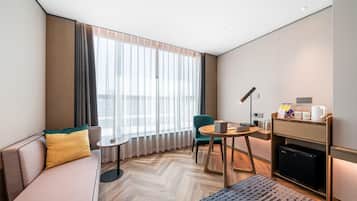 A modern hotel room with a sofa, a small table, a chair, a desk, a lamp, a coffee machine, and a mini fridge.