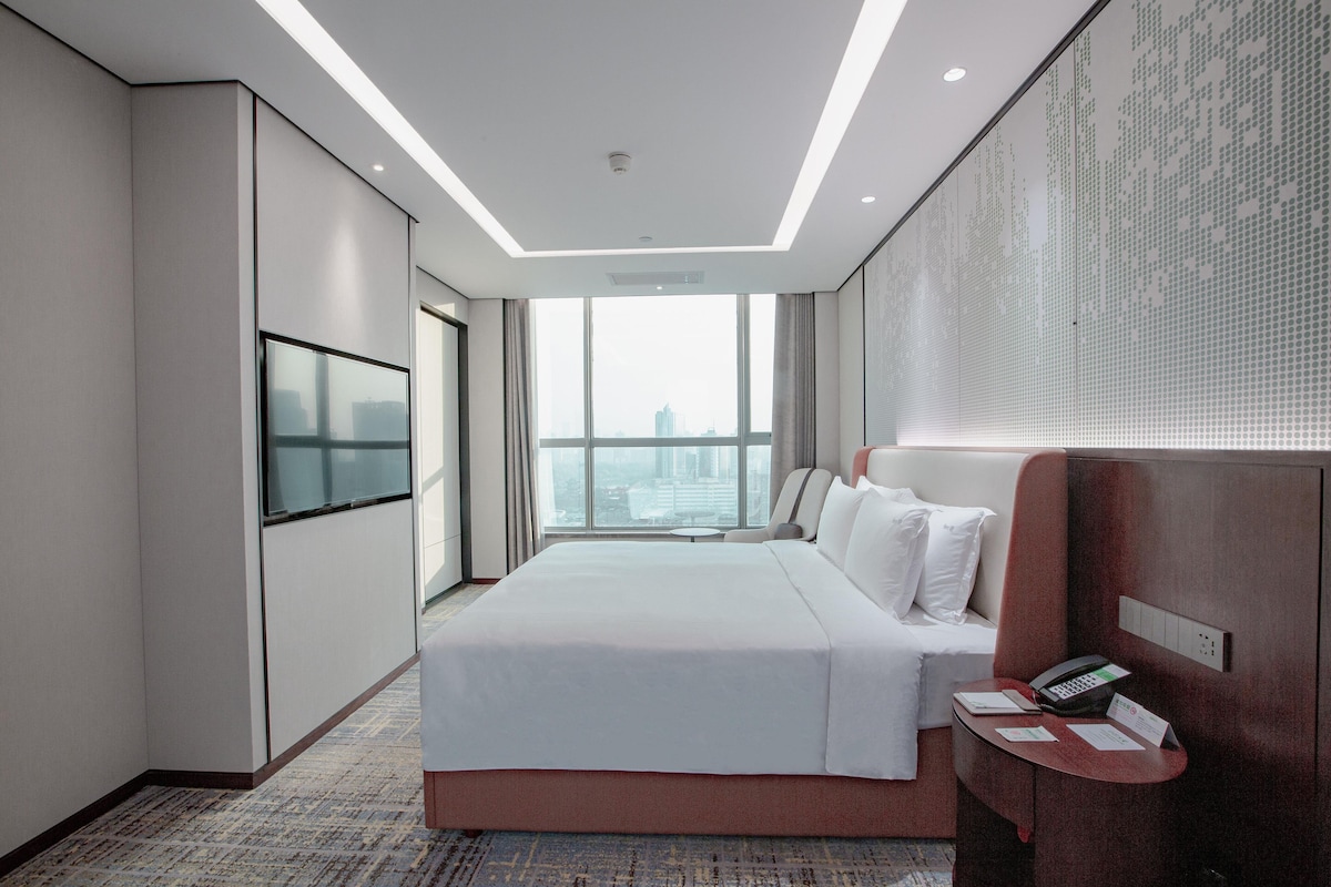 A hotel room with a large bed, a TV mounted on the wall, a desk with a phone, and a view of the city through large windows.