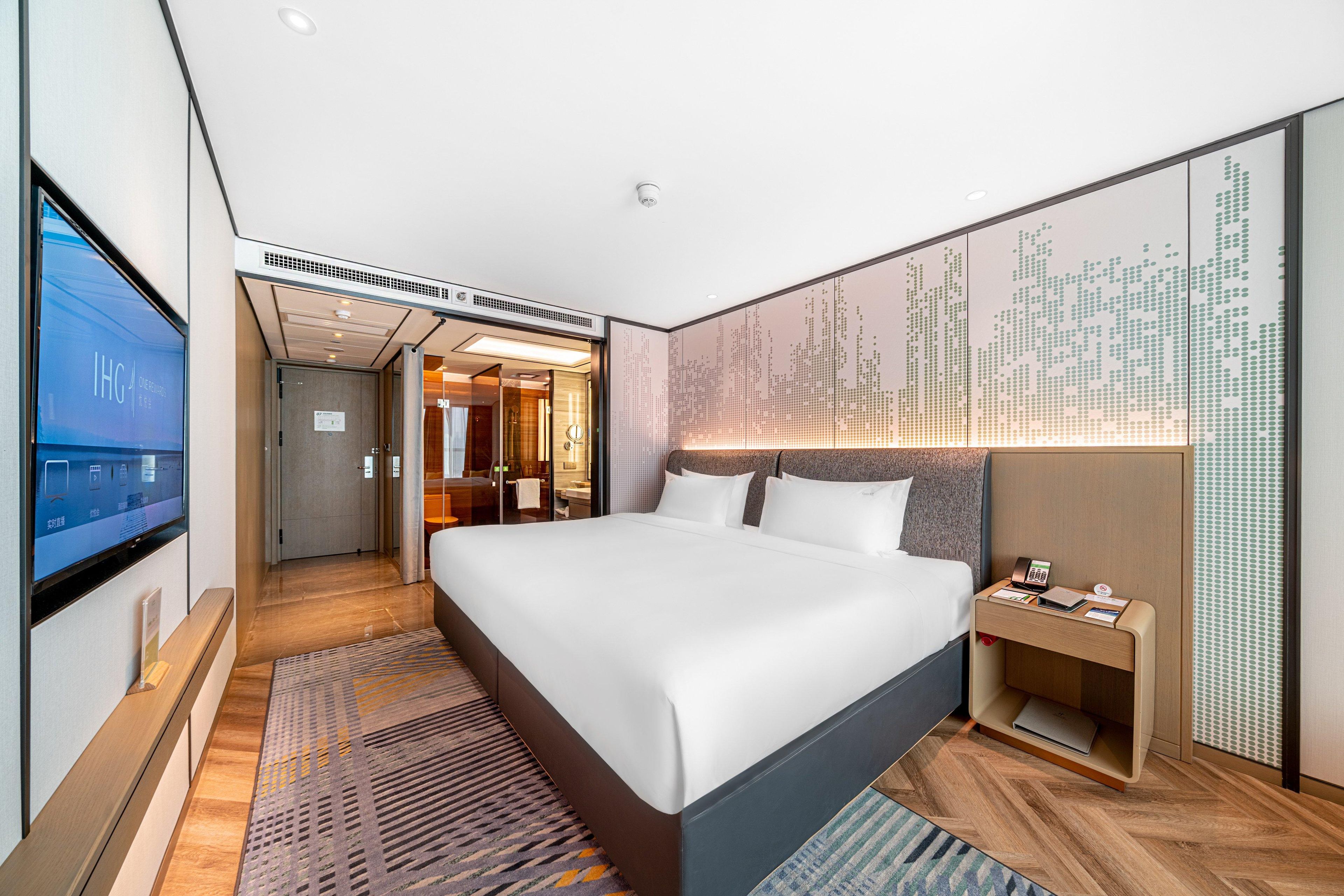 A modern hotel room with a large bed, a bedside table, a TV, and a wall-mounted artwork.