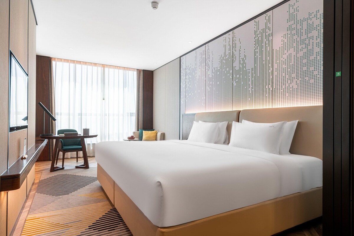 A hotel room with a large bed, a desk with a chair, a large window with curtains, and a wall with a digital display.