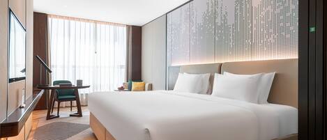 A hotel room with a large bed, a desk with a chair, a large window with curtains, and a wall with a digital display.