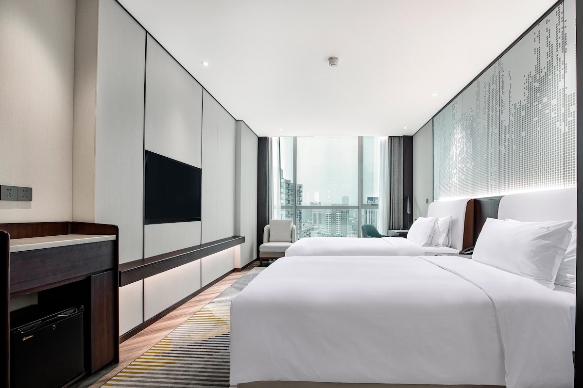 A modern hotel room with a large bed, a desk with a computer, a chair, and a city view.