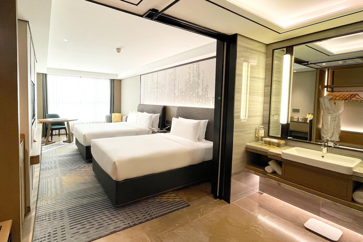 A hotel room with two beds, a desk, a chair, a large mirror, and a bathroom with a sink and towel rack.
