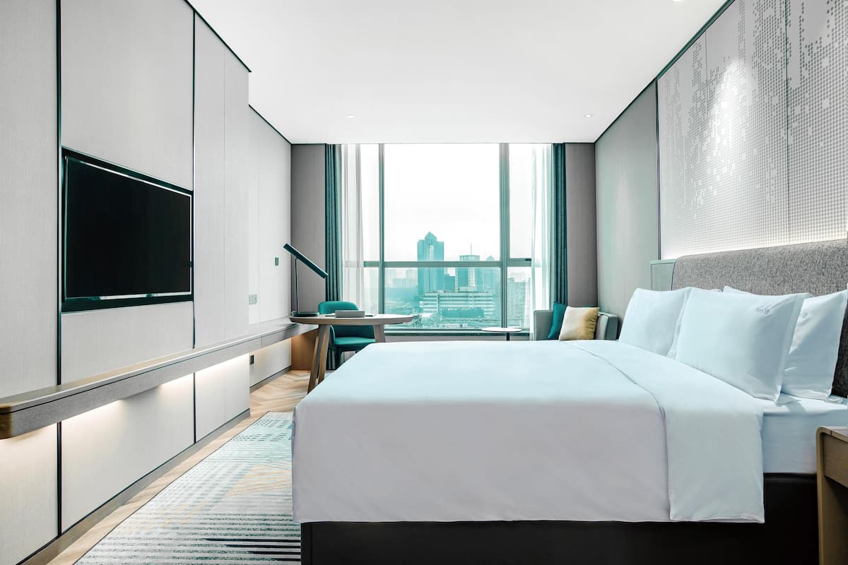 A modern hotel room with a large bed, a flat-screen TV, a desk, and a view of the cityscape.