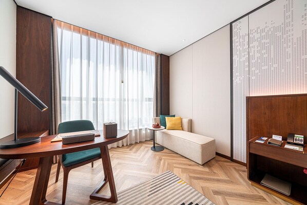 Una habitación de hotel moderna con piso de madera, una mesa redonda con lámpara, un sofá y un escritorio con computadora y teléfono.
