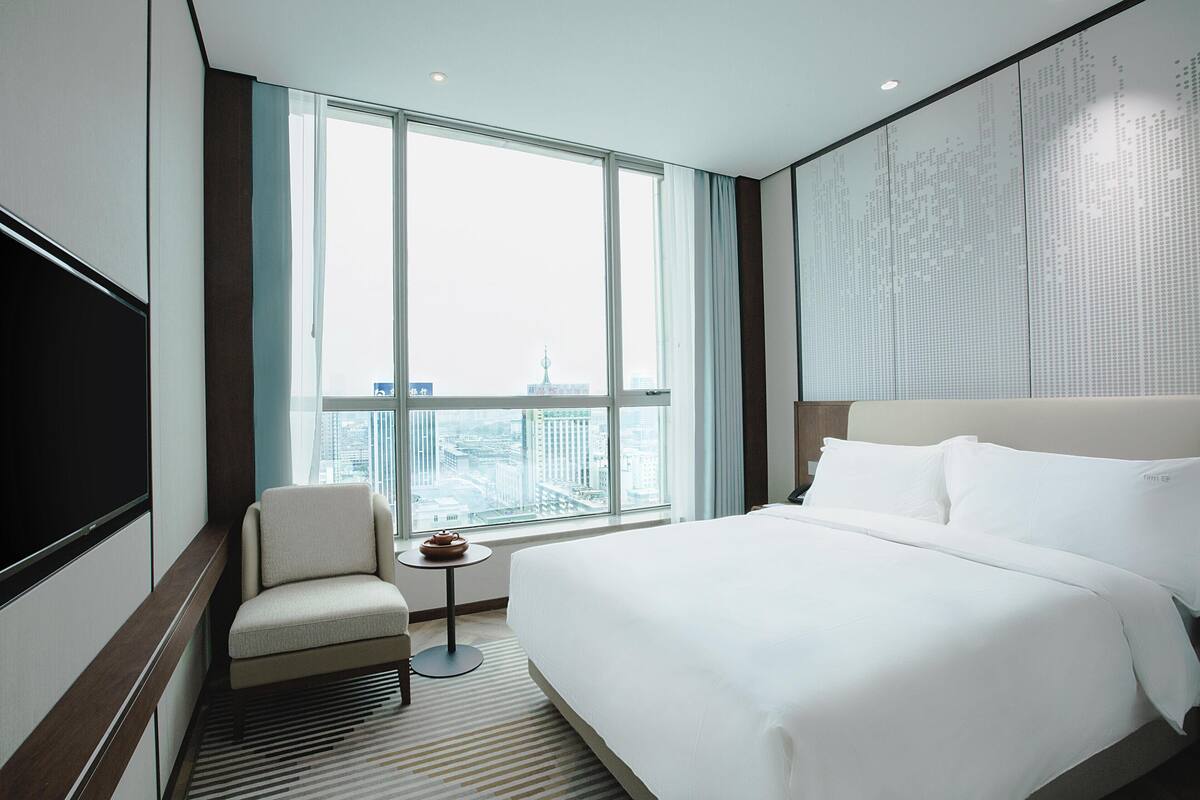A hotel room with a large bed, a chair, a small table, and a large window with a city view.