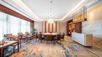 A modern hotel lobby with a round dining table, comfortable seating, a reception desk, and a large window with curtains.