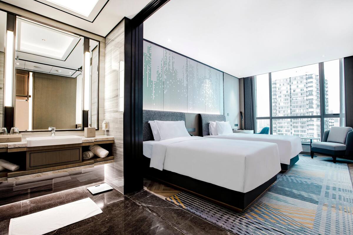A modern hotel room with a large bed, a bathroom with a mirror and sink, and a view of the city.