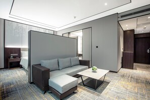 A modern hotel room with a sectional sofa, a small coffee table, a desk with a lamp, and a large window.