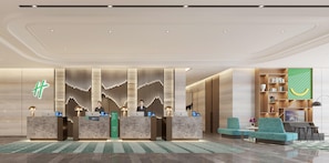 Zona de recepción de hotel con diseño moderno, que incluye un mostrador de recepción, dos empleados, un letrero verde y un área de descanso con un sofá y sillas.