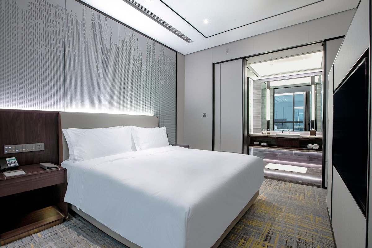 A modern hotel room with a large bed, a bedside table with a phone, a mirror, and a TV.