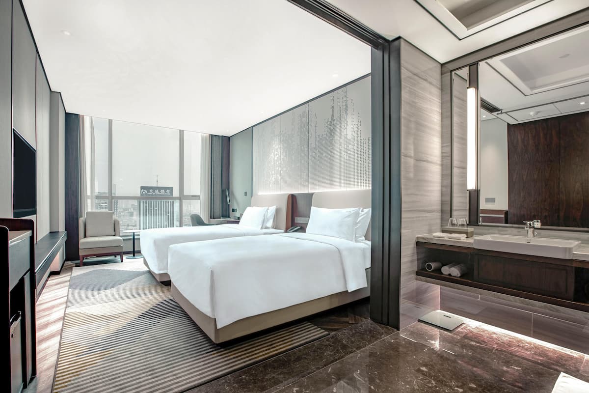 A modern hotel room with two beds, a large window, a bathroom with a mirror and sink, and a city view.