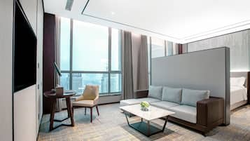 A modern hotel room with a large window, a sofa, a chair, a coffee table, and a TV.