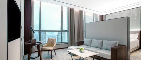 Una habitación de hotel moderna con un ventanal grande, un sofá, una silla, una mesita de centro y un televisor.