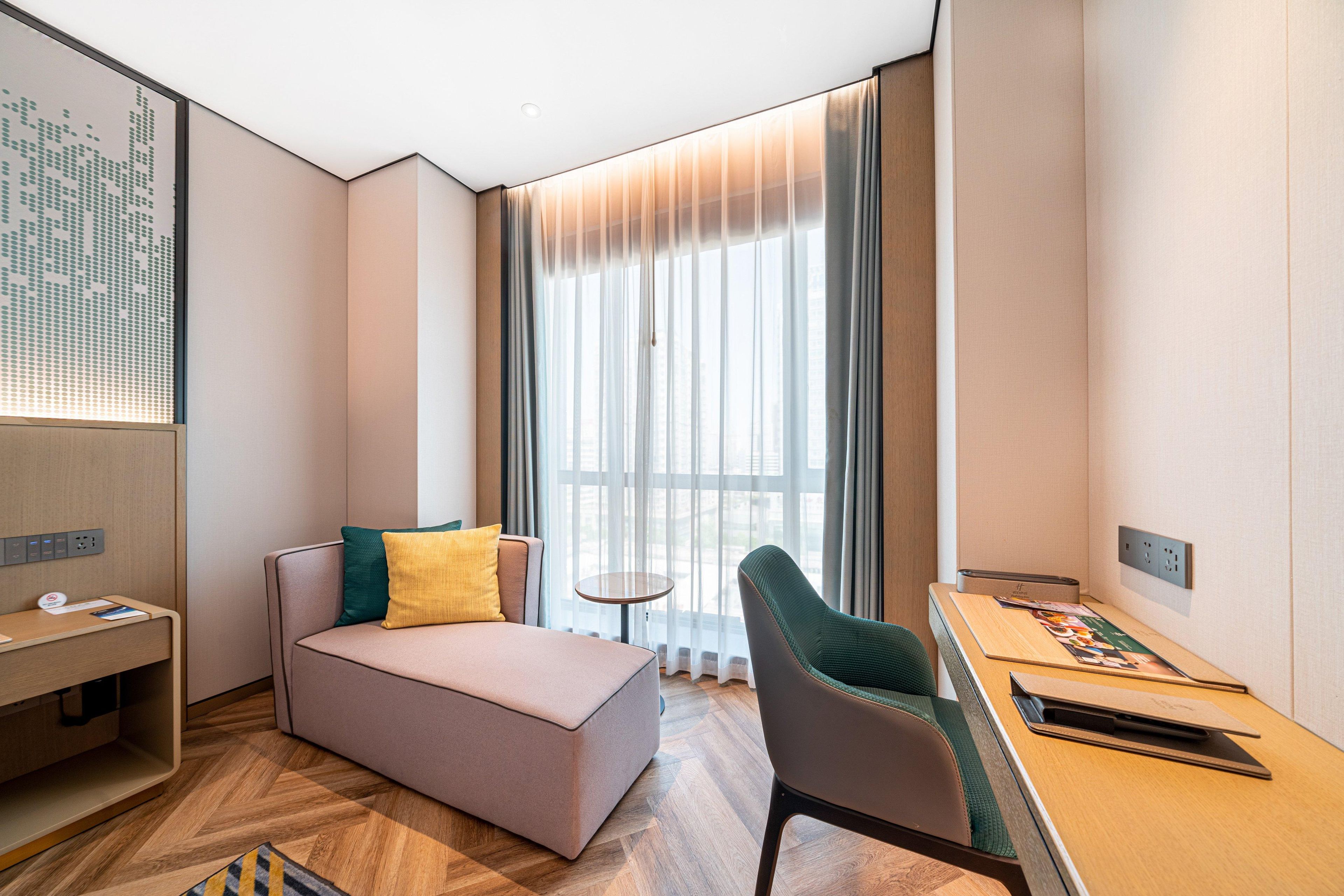Een moderne hotelkamer met een bank, een eettafeltje, een stoel en een groot raam met gordijnen.