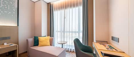 A modern hotel room with a sofa, a small table, a chair, and a large window with curtains.