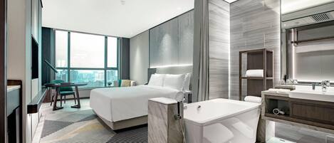 A modern hotel room with a large bed, a separate bathroom with a bathtub, a vanity, and a city view.