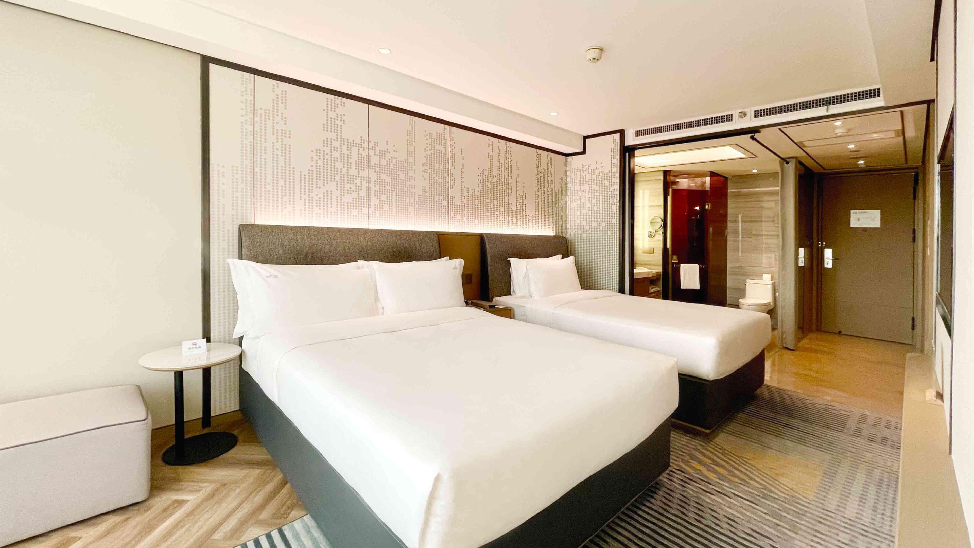 Ein Hotelzimmer mit zwei Betten, einem kleinen Nachttisch, einer runden Uhr und Blick ins Badezimmer.