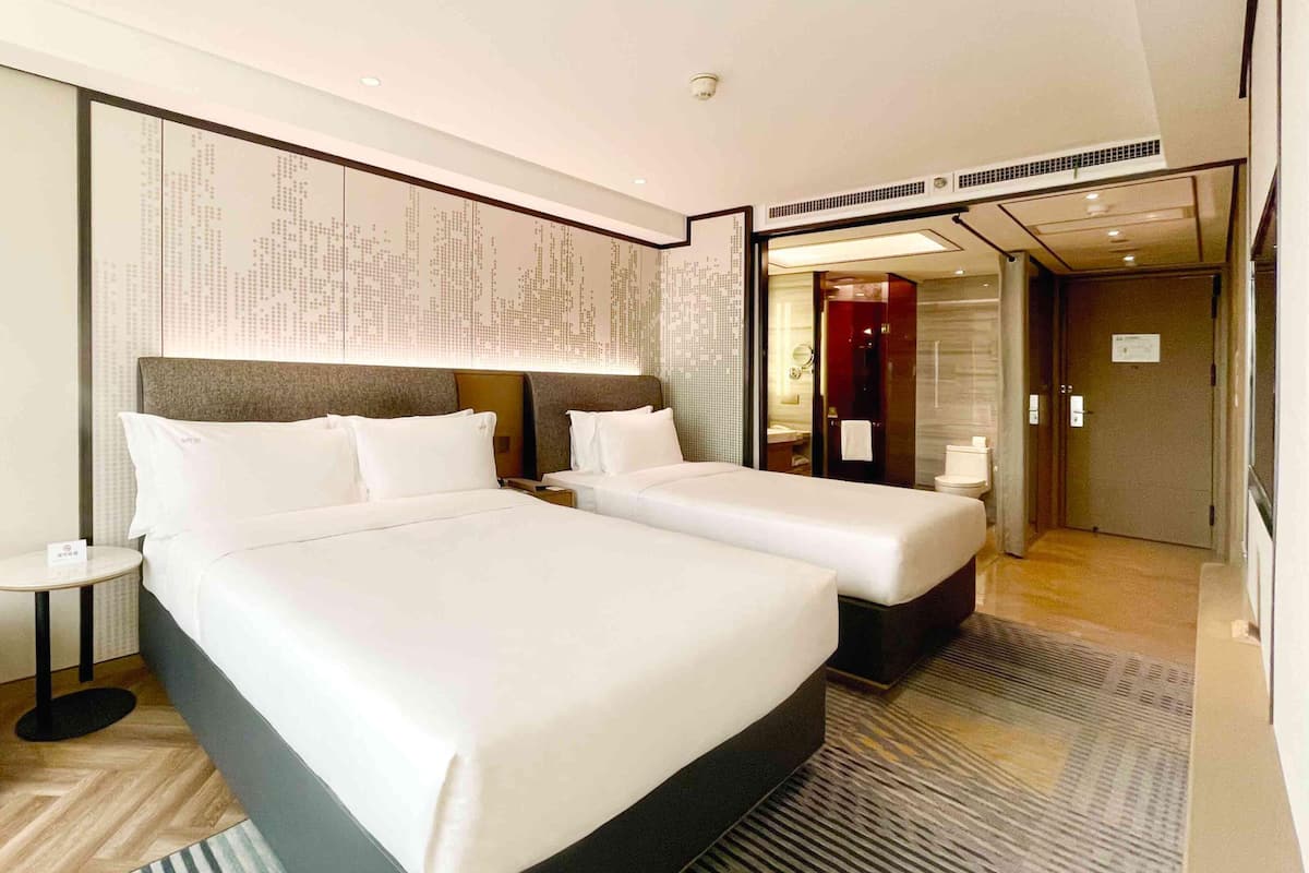 A hotel room with two beds, a small bedside table, a round clock, and a view of the bathroom.