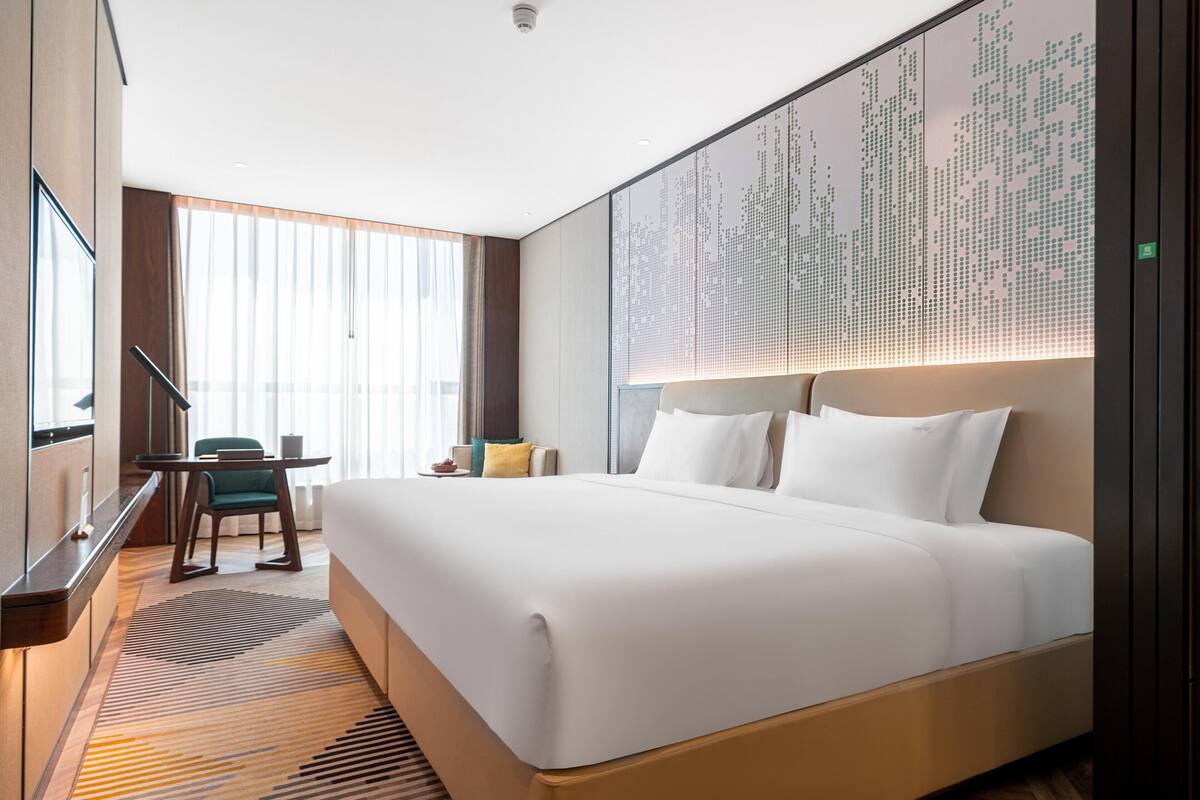 A hotel room with a large bed, a desk with a chair, a large window with curtains, and a wall with a digital display.
