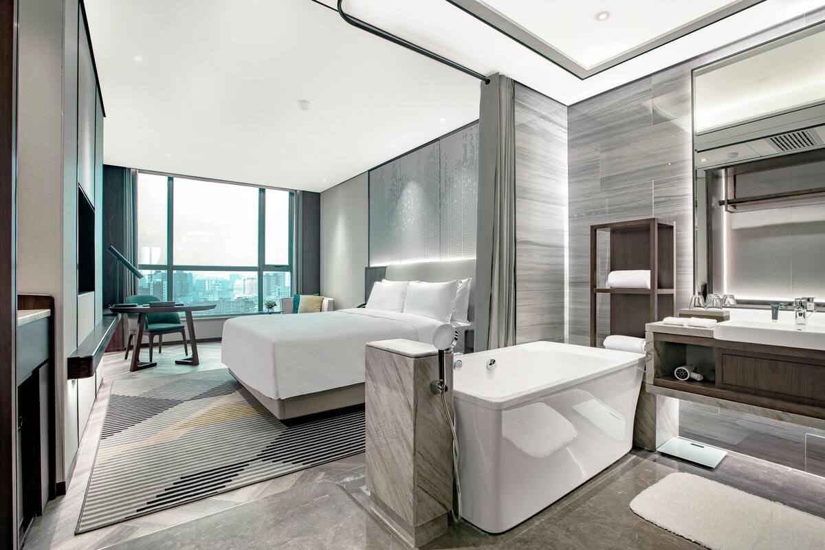 A modern hotel room with a large bed, a separate bathroom with a bathtub, a vanity, and a city view.