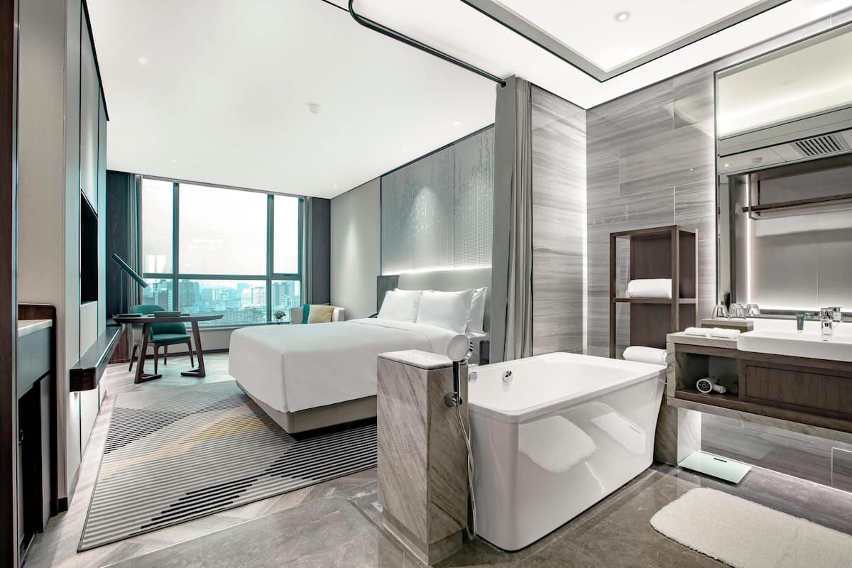 Ein modernes Hotelzimmer mit einem großen Bett, einem separaten Badezimmer mit Badewanne, einem Schminktisch und Blick auf die Stadt.