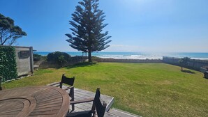 A backyard with a wooden deck, a table, and chairs facing a grassy area and a view of the ocean. - The Gin Trap (Pukehina)