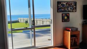 A room with a large sliding glass door leading to a wooden deck and a view of the ocean. There is a small wooden table with a mat, a shelf with a record player, and a framed picture on the wall. - The Gin Trap (Pukehina)